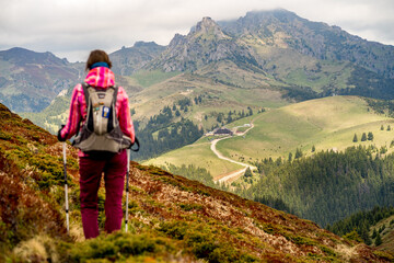 Dramatic landscape with a young mountain girl equipped with a backpack and hiking poles looking into the distance to the cottage at the base of the mountain.