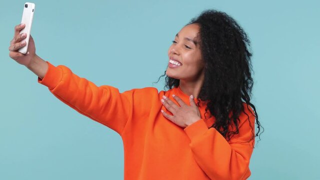 Fun cute young african american woman with curly hair lush hairstyle 20s years old wearing orange sweatshirt doing selfie shot on mobile phone isolated on pastel blue color background studio portrait