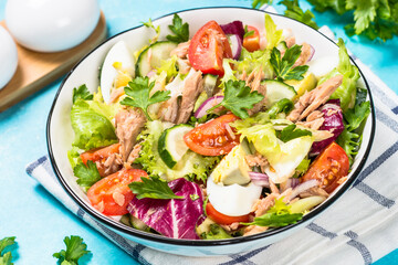 Tuna salad with green leaves, eggs and vegetables in white bowl at blue background.