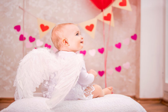 baby girl with white feather wings sits on white soft pillow among the Valentine's Day symbol, view from the back