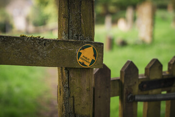 Sign: Public Footpath, seen on a wooden fence, with a blurry cemetery in the background, seen in Aysgarth, North Yorkshire, England, UK