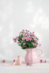 Pastel still life with pink flowers in a pink pitcher and silk ribbon on a white wooden table 