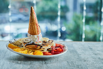 Pancakes with ice-cream cone, tangerine, strawberry, banana and chocolate syrup in the white plate on the marble background