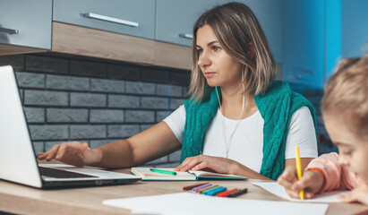 Caucasian mother is concentrated on her work at he laptop while daughter is drawing her her in the kitchen