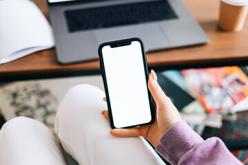 Woman holding a smartphone with a white screen mock up, resting on the armchair in living room at home.