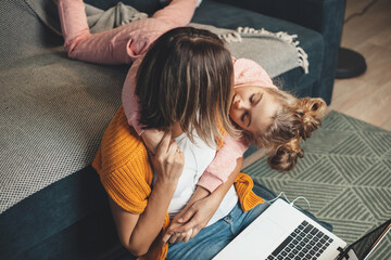 Caucasian mother is working on the floor using a laptop while her daughter is embracing her