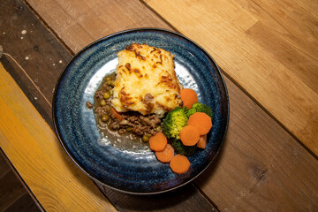 A delicious plate of Shepherds Pie with Mash Potato, Carrots and Broccoli on a wooden kitchen table
