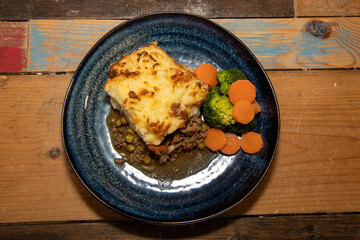 A delicious plate of Shepherds Pie with Mash Potato, Carrots and Broccoli on a wooden kitchen table