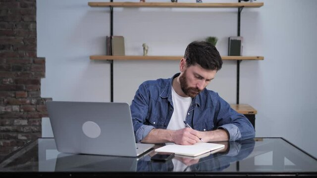 Close up view of young caucasian businessman in casual clothes occupied working from home using his laptop making notes in notebook looking concentrated focused serious. Work online connection.