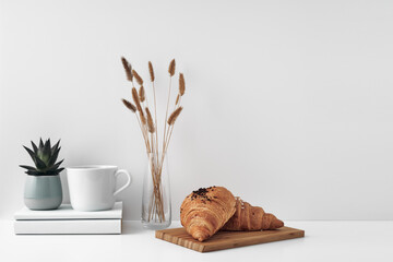 Croissant and a mug on a wooden stand, a vase and a pot with a plant on the books, white background. Eco-friendly materials in interior decor, minimalism. Copy space, mock up.