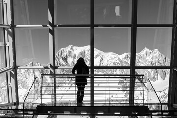 Woman from behind admiring Monte Bianco (Aosta Valley, Italy) behind a wall made of glass (Monte Bianco Skyway). Monochromatic