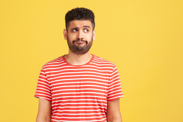 Doubtful bearded man in striped t-shirt standing with thoughtful expression, remembering or...