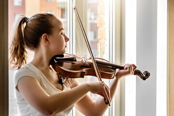 A young girl, a musician, plays the violin on the background of a window
