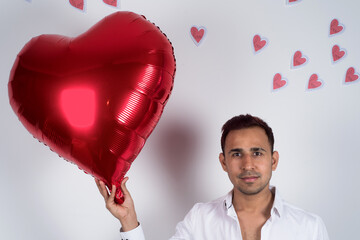Happy man, looking at the camera, holding a red heart balloon, heart papers flying behind him, white background. love and Valentine concept 