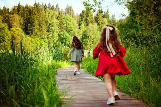 Two Little Girls With Long Hair In A Green And Red Dress Run Along A Wooden Path In The Park. A Fun Childhood Of The Two Sisters.