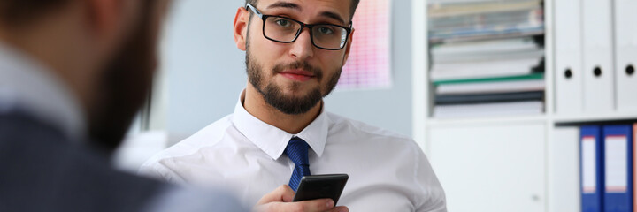 Bearded male worker using cellphone at workplace while chatting with his colleague in the office. Work and business concept