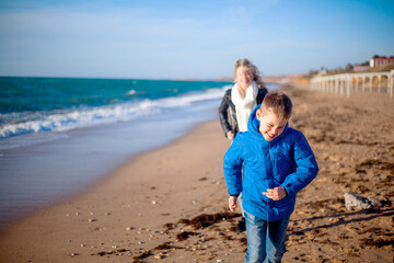 Happy family, mother with son walking wirh fun in the sea shore on windy day. People dressed warm clothes.