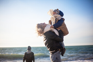 Happy family, mother with sons walking wirh fun in the sea shore on windy day. People dressed warm clothes.