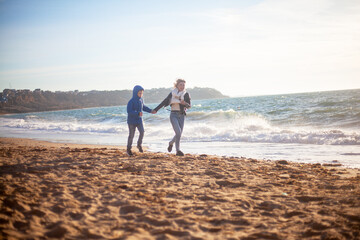 Happy family, mother with son walking wirh fun in the sea shore on windy day. People dressed warm clothes.