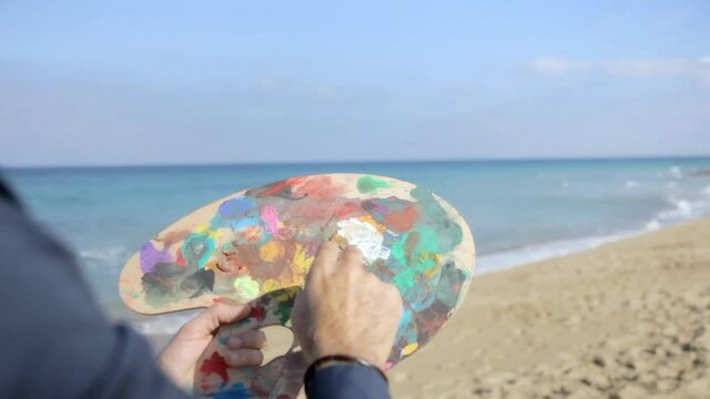 An inspired man (an artist), at a sandy beach, painting the sky with colors from a painter's palette. Creativity, inspiration. Handheld shot.
