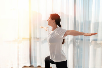 Yoga. A young woman trains at home, and smiles while doing a warrior pose. Sunlight from the window. The concept of home workouts