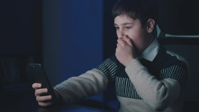 Afraid and shocked boy look at smartphone screen, cover mouth with hand at night at home. Schoolboy kid scared of bad news, read terrible message on mobile phone sitting at room. Watching horror video