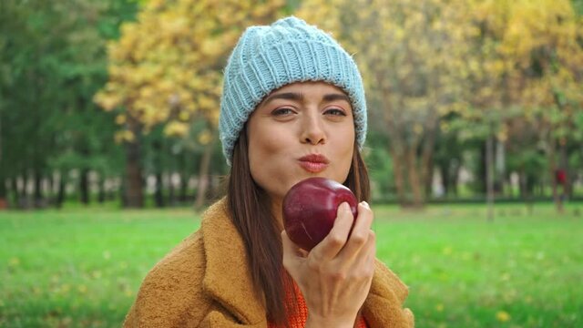 joyful woman eating juicy apple while looking at camera in park