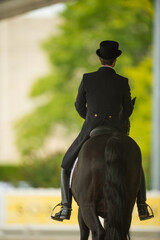 Dressage equestrian rider on black horse shot from behind dressed in dressage apparel attire black jacket with tails tall black boots beige jodhpurs at horse show  competition black leather saddle