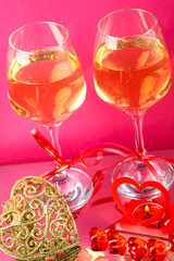 Two glasses with champagne tied with red ribbons on a pink background next to a souvenir heart and a candlestick heart with a burning candle.