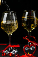Champagne is poured into glasses tied with a ribbon on a black background, next to a heart, a candlestick with a burning candle and a box with a ring.