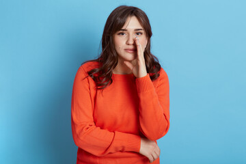 Young adorable European curious woman keeps hand near mouth and whispers secret, spreads rumors, dressed casually, isolated on blue background, telling interesting news.