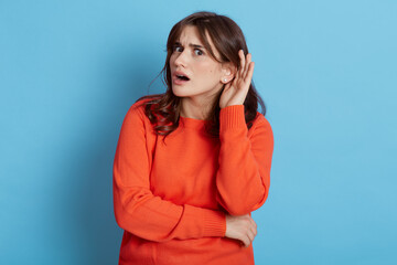 Attractive woman with dark hair listening to something interesting and holding hand near her ear, curious girl being attentive, keeps mouth opened, looks surprised, isolated over blue background.