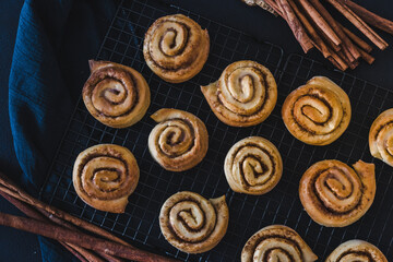 Homemade cinnamon rolls on a cooling rack with cinnamon sticks on dark blue background.