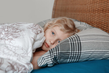 Pensive sad little Caucasian blonde child girl lying on pillow in bed at home. Child lying with open eyes thinking, dreaming. Candid authentic home life. Sweet dreams and bed vibes.