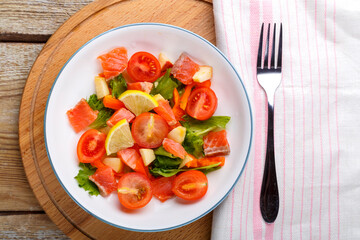 Salad with salmon and cherry tomatoes and green salad in a plate on a wooden table on a wooden stand with a fork and napkin next to it.