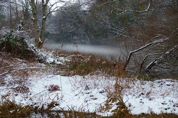 scenic winter view across the river with snow covered banks to a pond with mist over the water