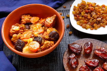 Jewish cuisine dish sweet zimmes with dates, carrots and turkey meat in a brown plate on a black board on a blue background near raisins and dates.