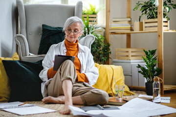 Portrait of senior woman architect sitting on floor indoors at home, working.