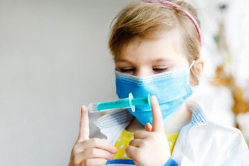 Little girl makes injection. Cute child with medical mask playing doctor, holding syringe with vaccine. Coronavirus covid vaccination concept. Kid play role game at home.