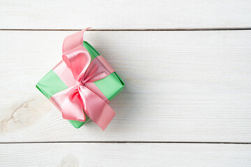 A small gift box in green packaging with a pink ribbon on a white wooden background. Top view with copy space. The concept of a Festive background.