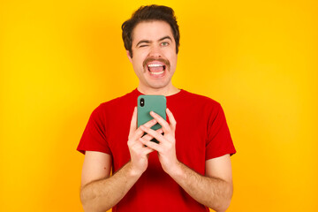 Young Caucasian man wearing red t-shirt standing against yellow background taking a selfie  celebrating success