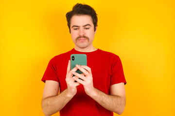 Excited Young Caucasian man wearing red t-shirt standing against yellow background  winking and eye hold smart phone use read social network news