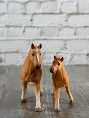 Two brown toy horses, a mother and a foal, against a light brick wall.