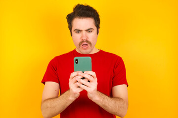 Focused Young Caucasian man wearing red t-shirt standing against yellow background use smartphone reading social media news, or important e-mail