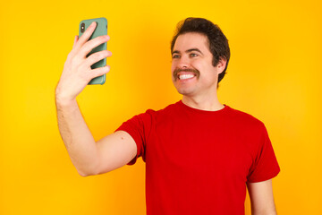 Young Caucasian man wearing red t-shirt standing against yellow background smiling and taking a selfie ready to post it on her social media.