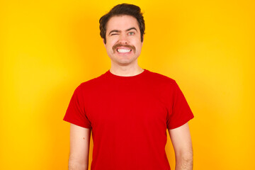 Fototapeta premium Coquettish Young Caucasian man wearing red t-shirt standing against yellow background smiling happily, blinking at camera in a playful manner, flirting with you.