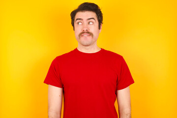 Obraz premium Amazed Young Caucasian man wearing red t-shirt standing against yellow background bitting lip and looking tricky to empty space.