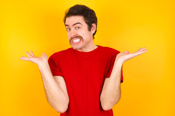 Clueless Young Caucasian man wearing red t-shirt standing against yellow background shrugs...