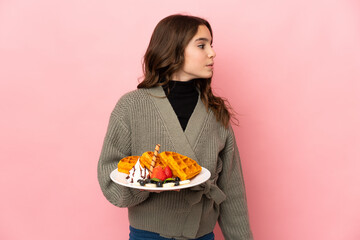 Little girl holding waffles isolated on pink background looking to the side