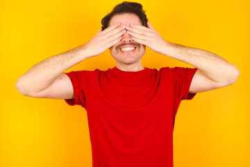 Fototapeta na wymiar Young Caucasian man wearing red t-shirt standing against yellow background covering eyes with hands smiling cheerful and funny. Blind concept.